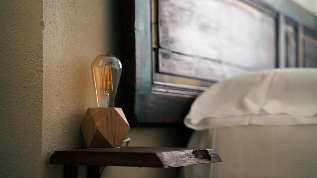 A bedside table with a light bulb and a wooden object, a white bed, and a wall with a textured surface.