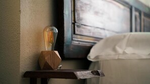 A bedside table with a light bulb and a wooden object, a white bed, and a wall with a textured surface. - Piccolo Borgo (Chiavari)