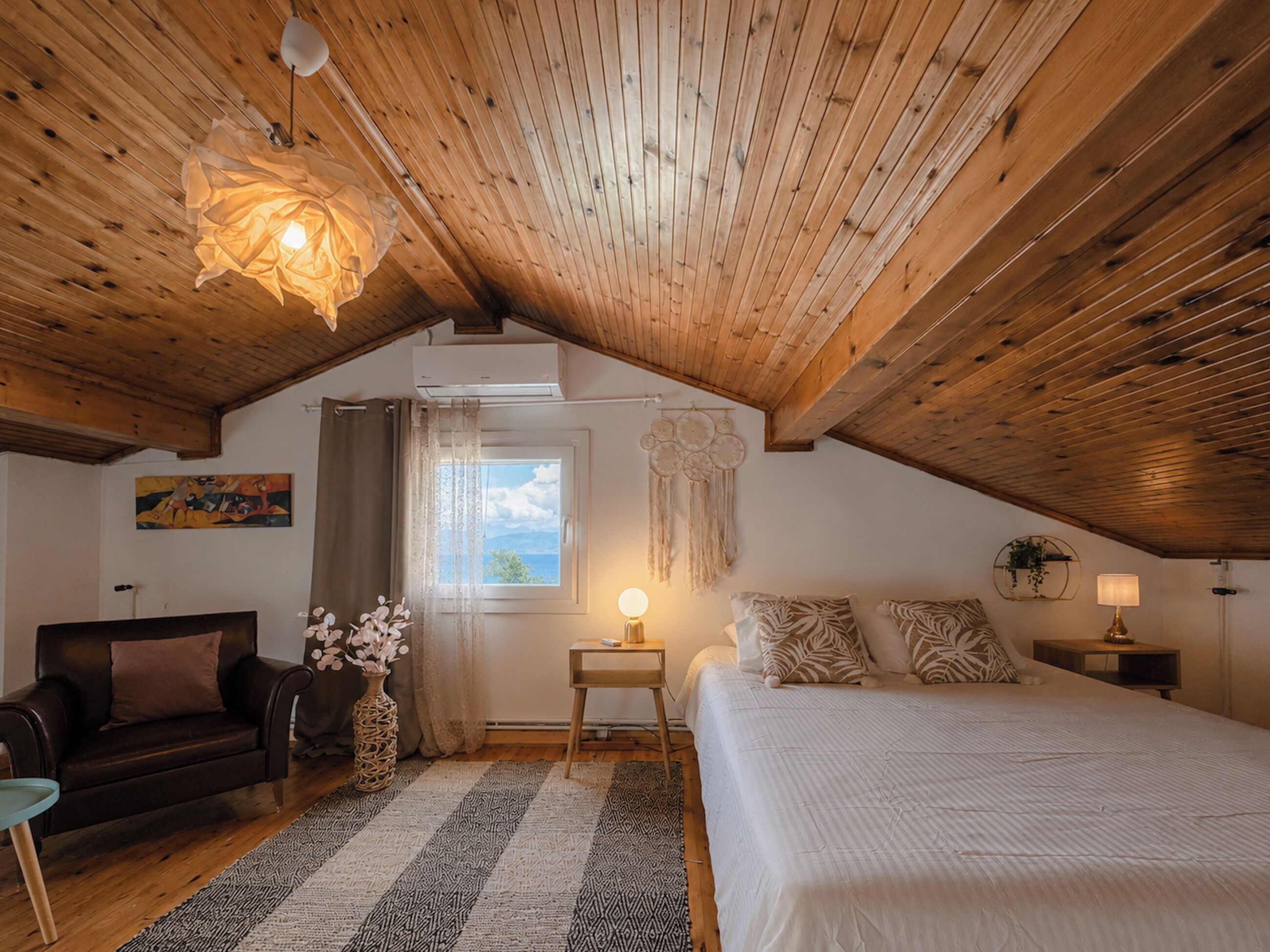 Une chambre confortable avec un plafond en pente, un lit, une table de chevet, une lampe, une chaise et une fenêtre donnant sur un beau paysage.