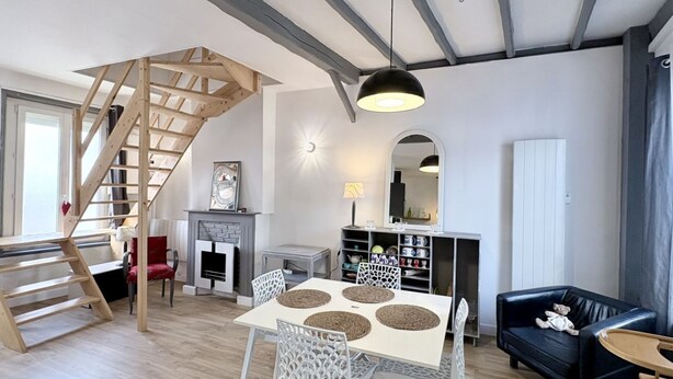 Ein modernes Wohnzimmer mit einer Holztreppe, einem Kamin, einem Esstisch mit Stühlen, einem schwarzen Ledersofa und einer Decke mit freiliegenden Balken.