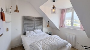 Ein Schlafzimmer mit schrägverdeckter Decke, einem Bett mit weißer Bettwäsche, einem Heizkörper und einem Fenster mit Vorhängen.