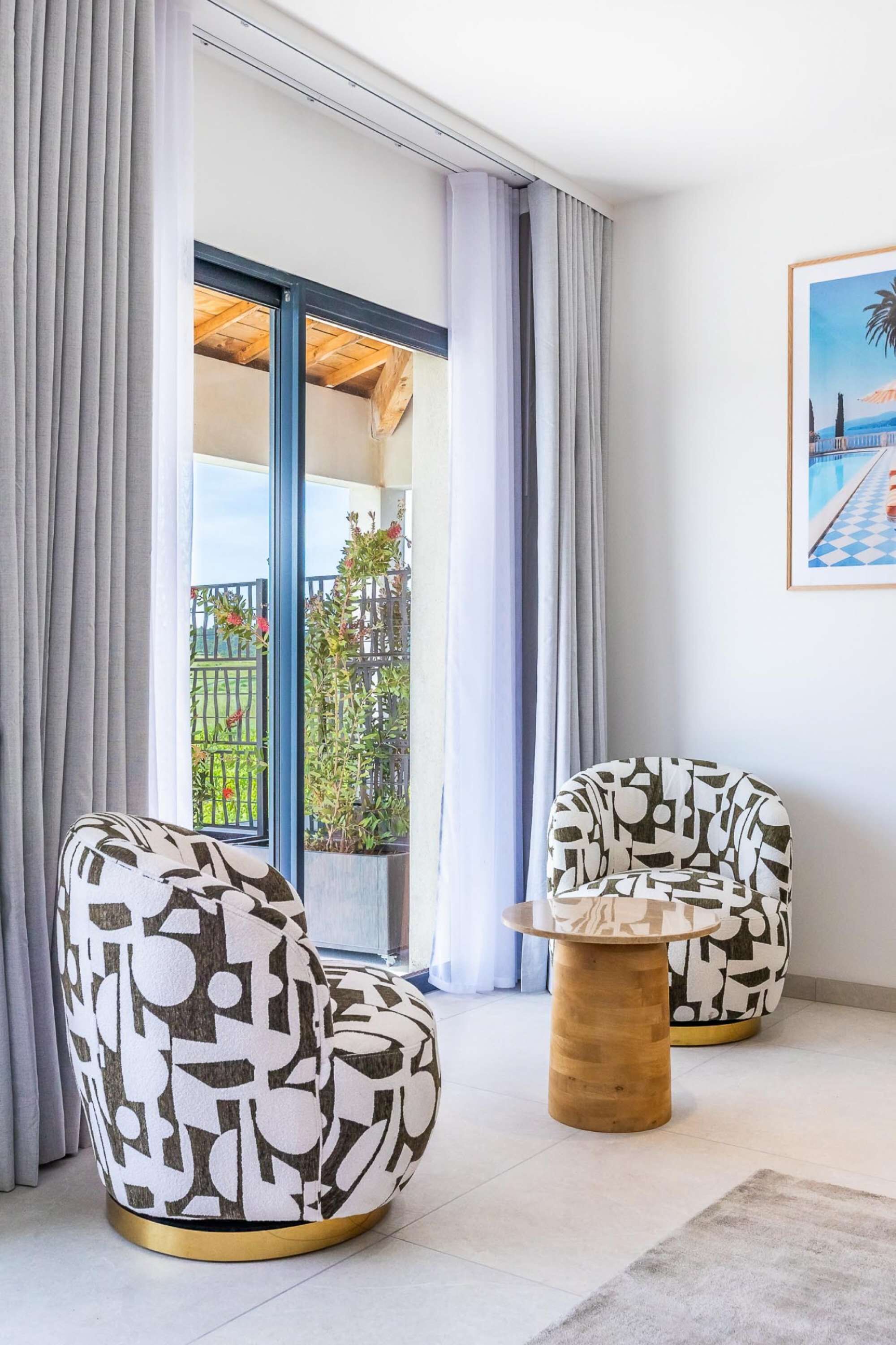 Een moderne kamer met twee gestoffeerde fauteuils, een houten bijzettafeltje en uitzicht op een balkon met planten en een hek.