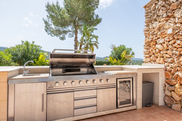Une cuisine extérieure équipée d’un barbecue, d’un évier et de tiroirs de rangement, adossée à un mur en pierre et entourée de verdure.