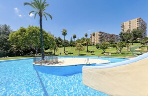 A swimming pool with a palm tree, a grassy area, and a multi-story building in the background. - ANDREA 2 - Modern studio apartment with beautiful view in Ju (Arroyo de la Miel)