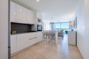 A modern kitchen with white cabinets, a central island with bar stools, and a view of the outdoors through large windows. - VIOLETA CB - Modern apartment with a beautiful view (La Cala de Mijas)