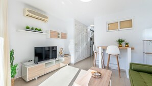 Un salon moderne avec un téléviseur, des meubles blancs, un coin repas et une cuisine en arrière-plan.