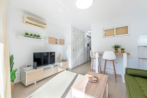Ein modernes Wohnzimmer mit Fernseher, weißen Möbeln, einem Essbereich und einer Küche im Hintergrund.