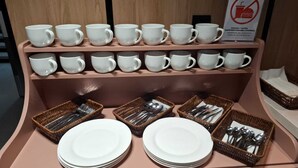 Une étagère présente une collection de tasses et d’assiettes blanches, accompagnée de paniers remplis de couverts.