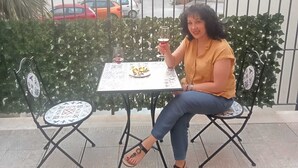 Uma pessoa sentada à mesa com um prato de comida e uma bebida, com uma cerca de metal e vegetação ao fundo.