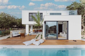 Ett modernt hus med en swimmingpool, utomhusmöbler och en glasdörr som leder in till ett vardagsrum.