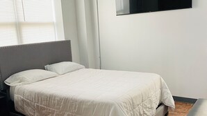 Une chambre à coucher comprenant un lit, un banc, une fenêtre avec des stores et un téléviseur fixé au mur.