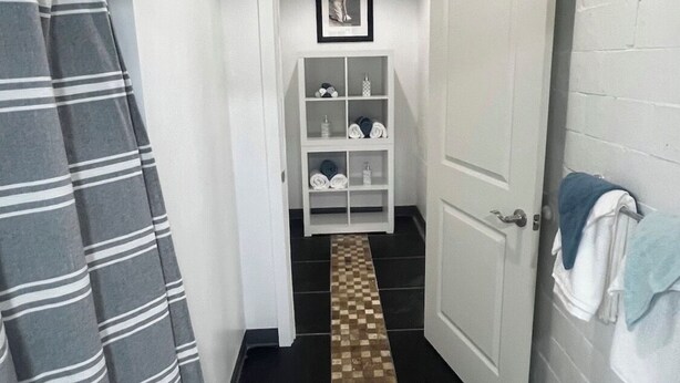 Un couloir étroit, doté d’un sol à carreaux, mène à une porte blanche, à un rideau de douche à rayures et à une étagère où sont pliées des serviettes.