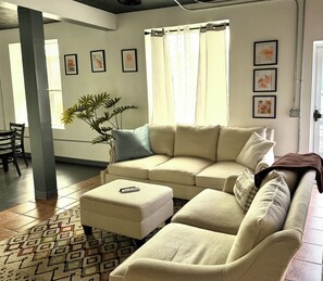 Un salon avec un canapé beige, un pouf assorti et une plante en pot.