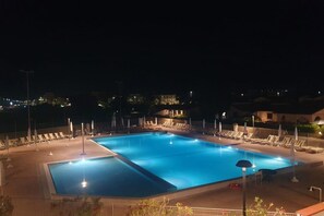 Una piscina illuminata di notte con lettini e ombrelloni, circondata da edifici.