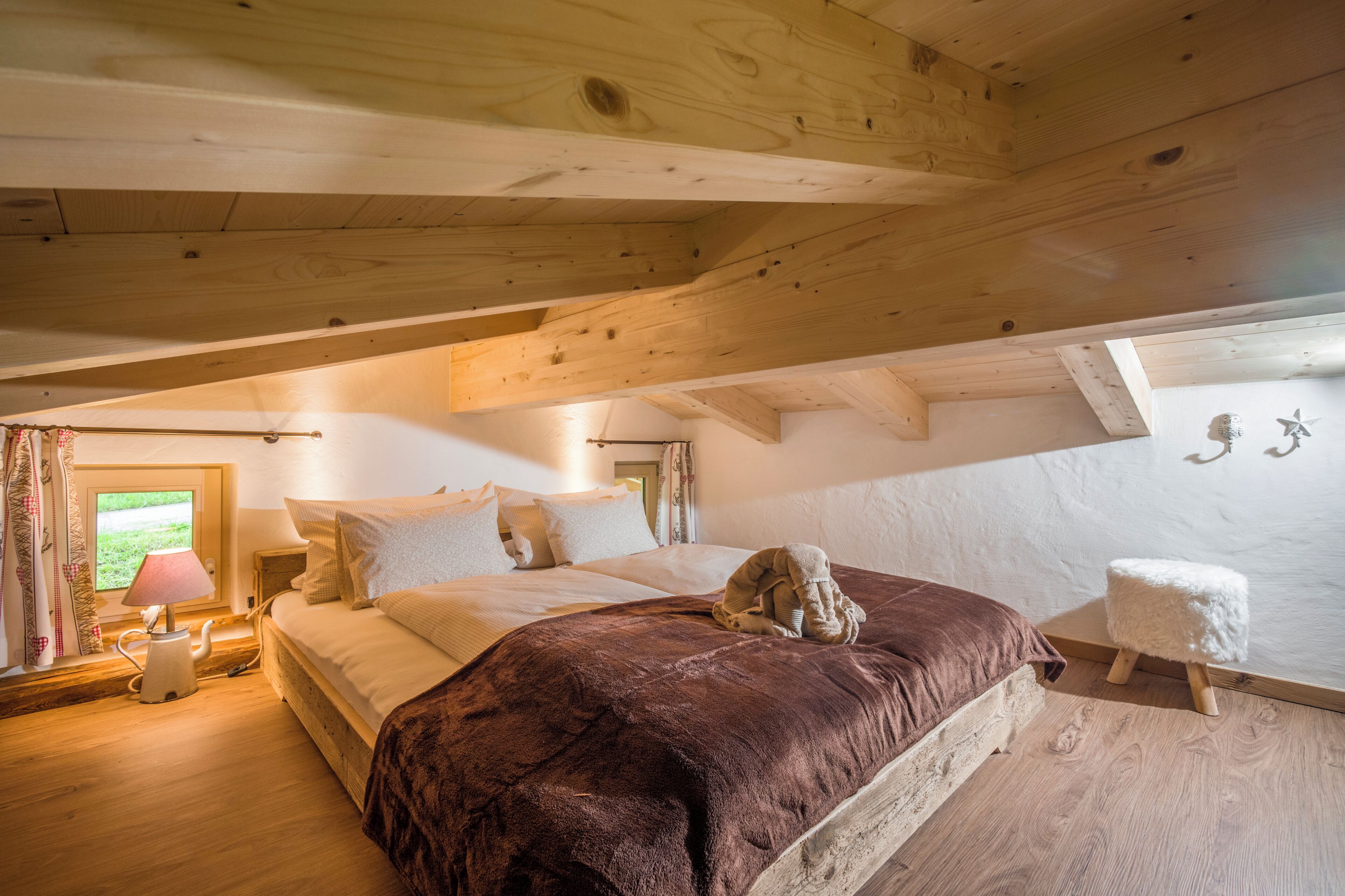 A bedroom with a wooden ceiling, a bed with a brown blanket, a nightstand with a lamp, and a window with curtains.