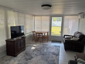 A living room with a TV, wooden table, chairs, a leather couch, and a door leading outside. - Charming 2-bedroom house in beautiful Clearwater with AC washer dryer (Clearwater)