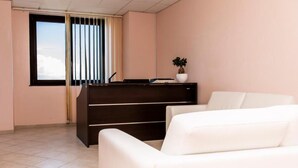 Een kamer met een piano, een witte bank, een raam met zonwering en een potplant.