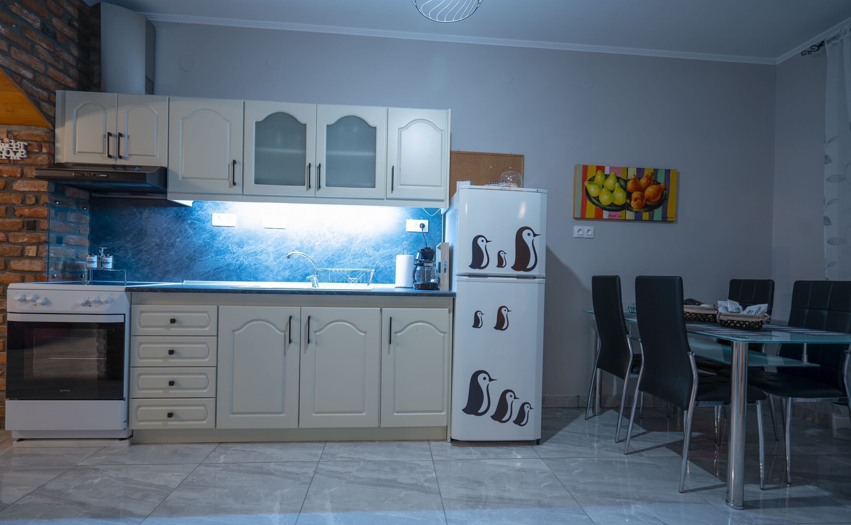 Une cuisine avec des meubles blancs, un réfrigérateur décoré d’autocollants représentant des pingouins, un coin repas avec une table et des chaises en verre, et une peinture de fruits accrochée au mur.