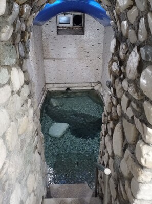 A narrow stone-walled passage with a small window and a metal grate, leading to a water-filled area with a single ice block. - To allo spiti Aridea (Aridea)