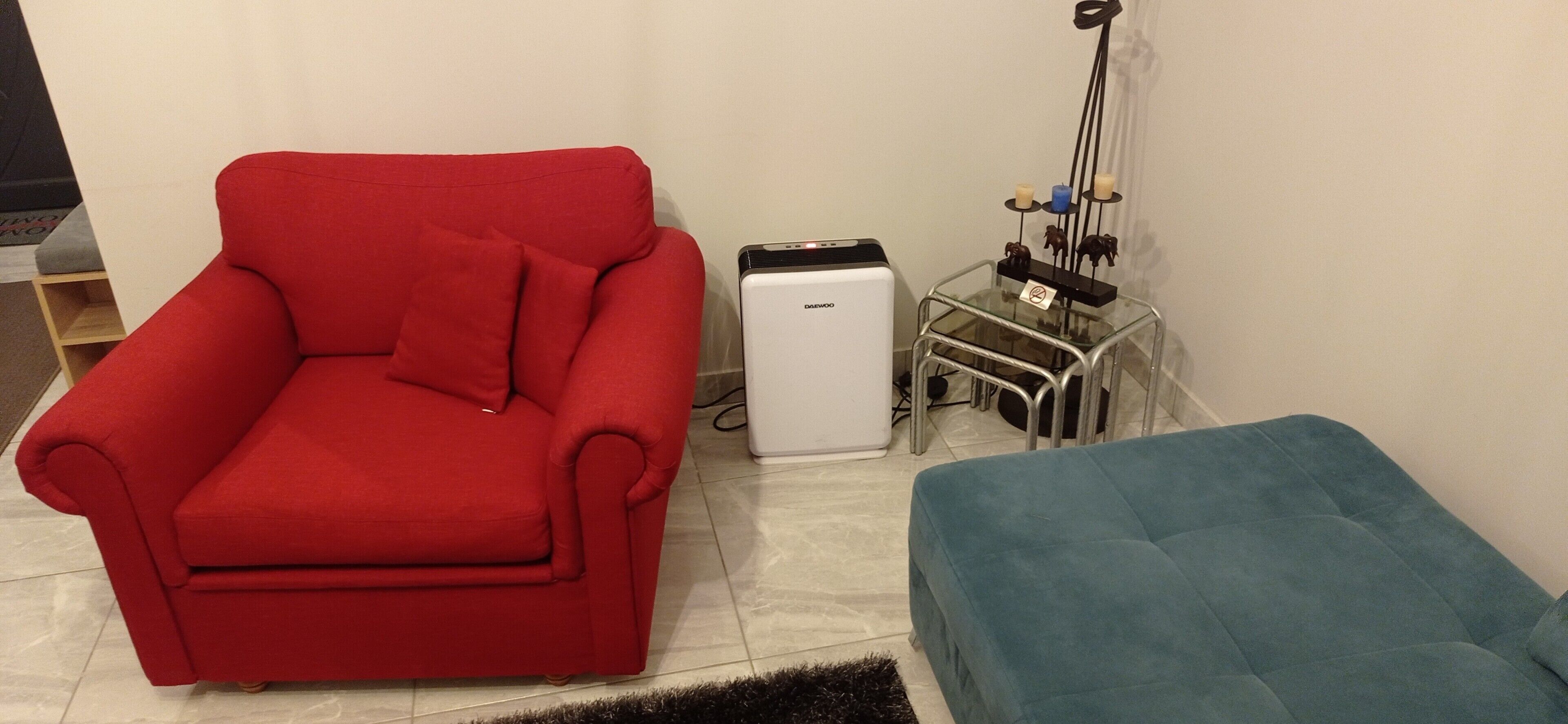 Een rode fauteuil, een klein wit apparaat en een blauwe poef in een kamer met betegelde vloer.