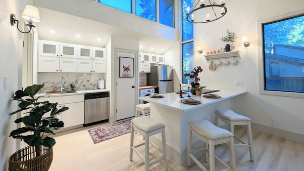 A modern kitchen with white cabinets, a central island with stools, a chandelier, and large windows. - Tamarron Treetop | AC, Pool, Hot Tub, Golf & Ski (Durango)