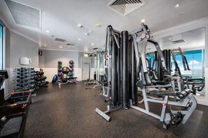 A gym with various exercise equipment, a wall-mounted rack of dumbbells, and a large window providing a view. - Iconic QV Aparment Hideaway | Chic 2BR CBD Escape (Melbourne)