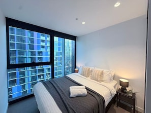 A modern bedroom with a large window, a bed with white linens, a bedside table with a lamp, and a view of a cityscape. - 2BR/2BA Urban Sanctuary: Views from 33rd Flr (Melbourne)