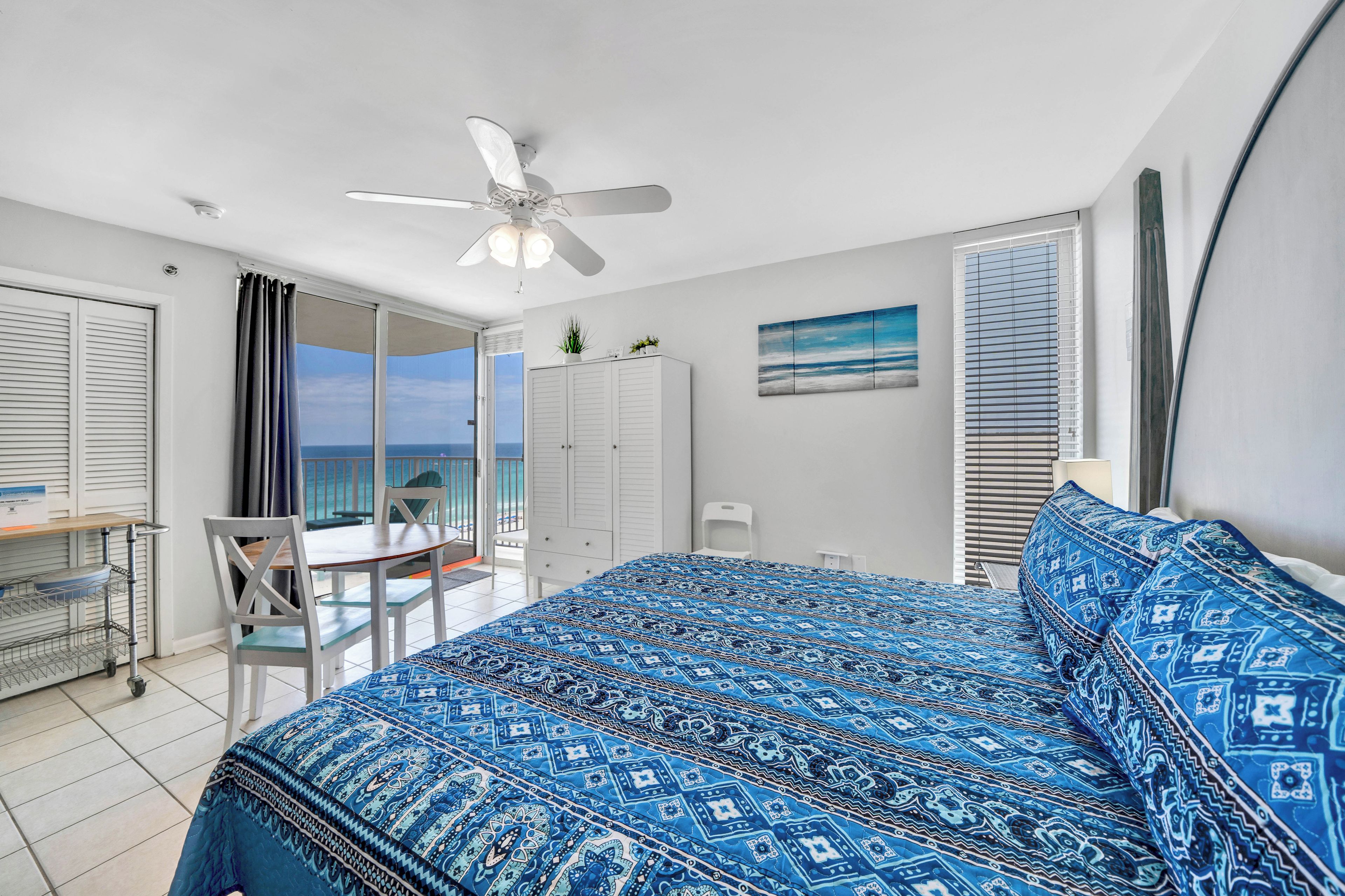 Ein Schlafzimmer mit einem Bett, einem Deckenventilator, einem Schreibtisch mit Stuhl und Meerblick durch eine Balkontür.