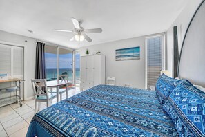 Ein Schlafzimmer mit einem Bett, einem Deckenventilator, einem Schreibtisch mit Stuhl und Meerblick durch eine Balkontür.