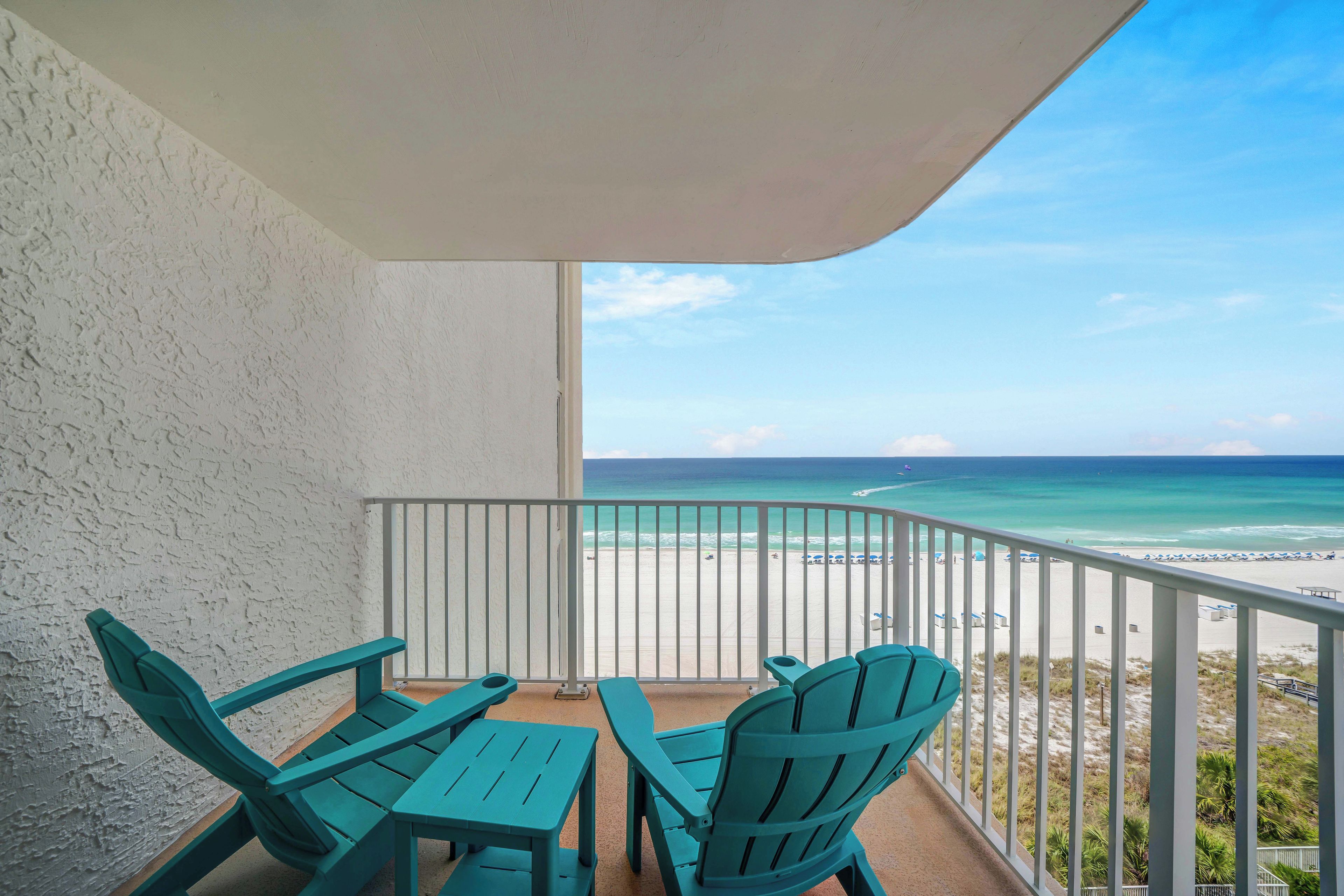 Un balcone con due poltrone Adirondack verdi e un tavolo, con vista su una spiaggia e acque turchesi cristalline e una riva sabbiosa.