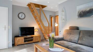 Een woonkamer met een houten tv-meubel, een bank, een bijzettafeltje met een vaas, een houten trap en een klok aan de muur.