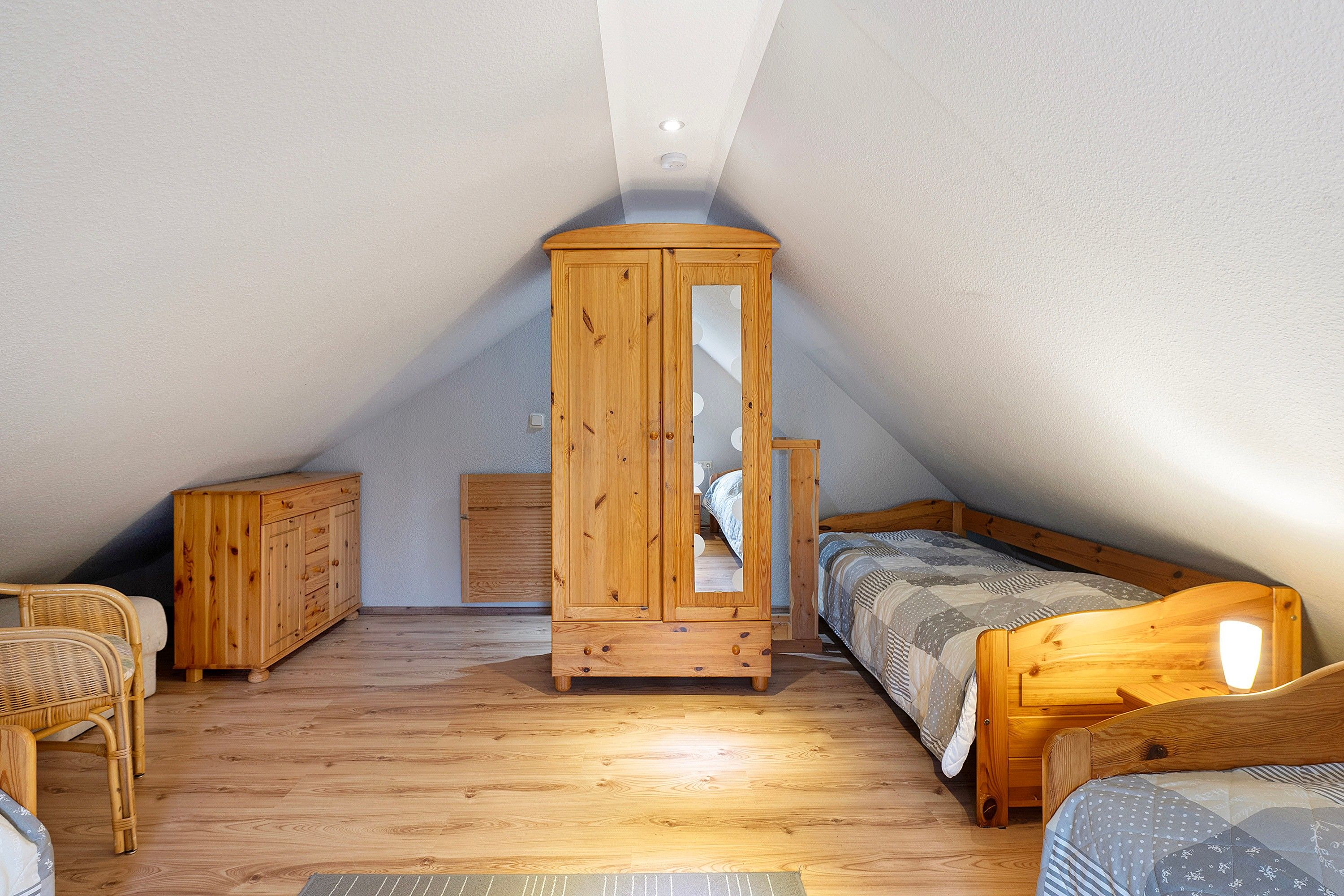 A small attic bedroom with a sloped ceiling, wooden wardrobe, two single beds, a wicker chair, and a small bedside table.