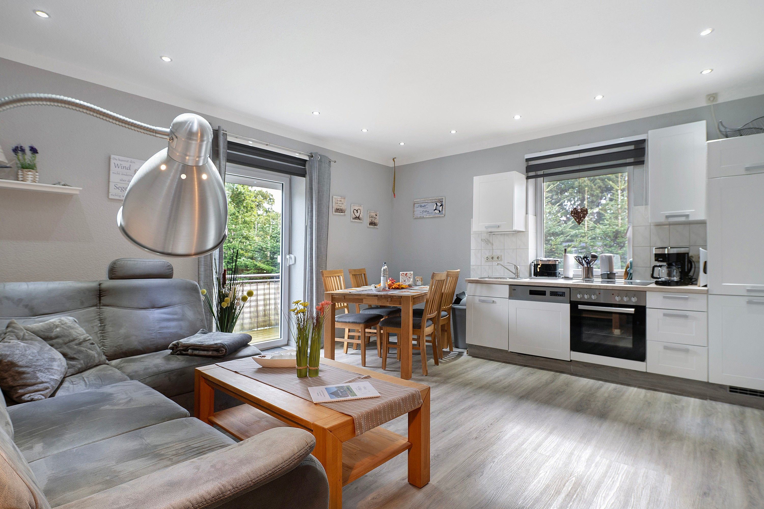 Eine moderne Küche mit weißen Schränken, ein Essbereich mit einem Holztisch und -stühlen sowie ein graues Sofa in einem Wohnzimmer mit einem großen Fenster.
