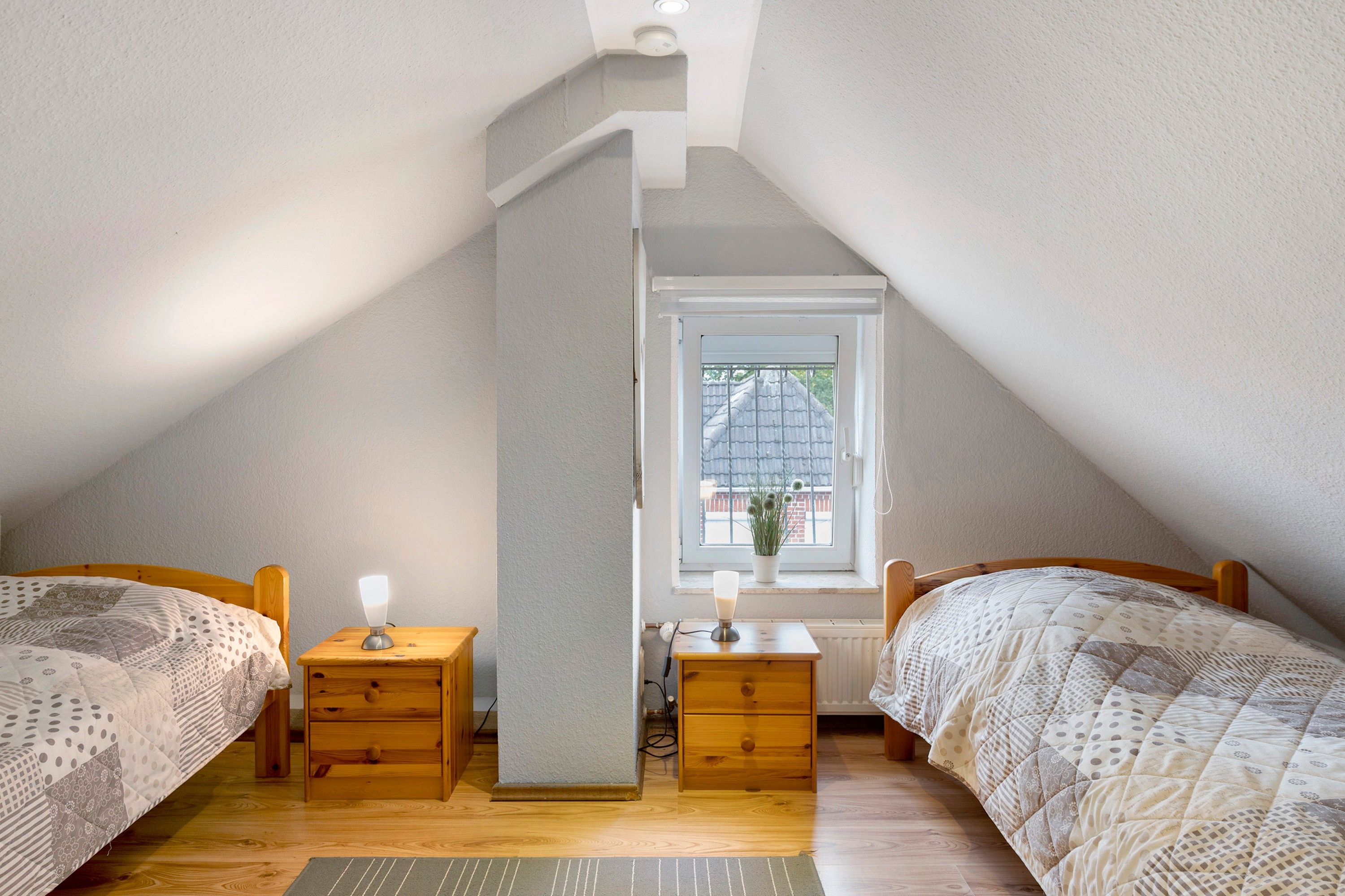 A bedroom with two wooden beds, a nightstand with a lamp, a dresser, and a window with a view of a house.