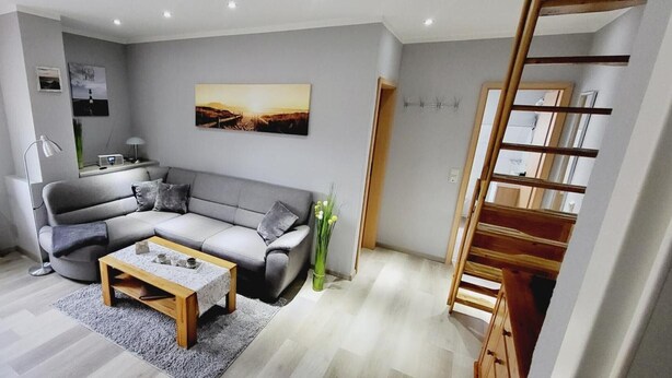 A modern living room with a grey sofa, a wooden coffee table, a radiator, a window with blinds, a painting on the wall, and a wooden staircase.