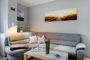 Un salon avec un canapé gris, une petite table sur laquelle est posé un vase de fleurs et, au mur, un tableau représentant un coucher de soleil.