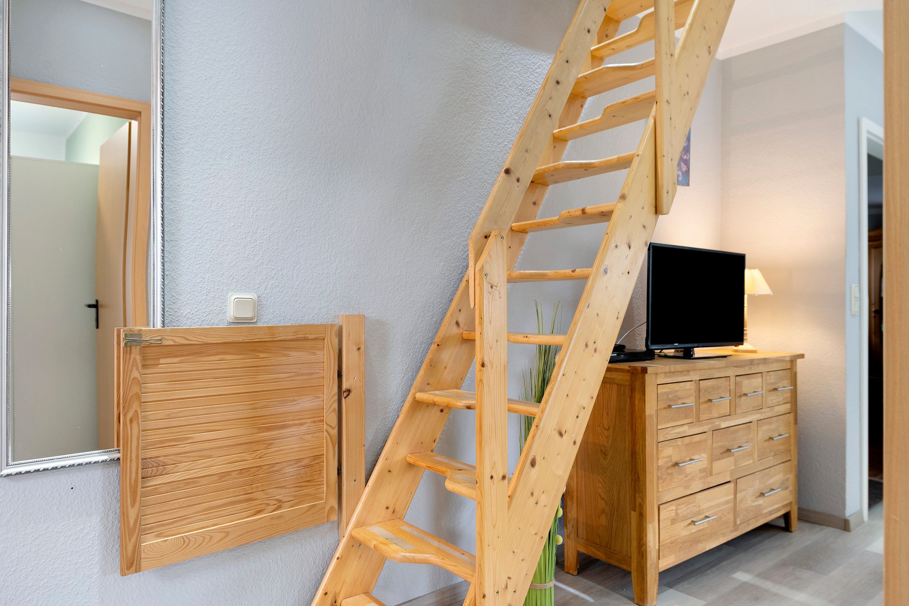 Eine Holzleiter, die an einer Wand lehnt, ein Fernseher auf einer Holzkommode und eine Tür mit einem Spiegel.
