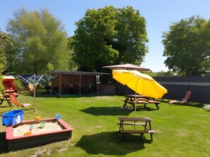 Un jardin avec une table de pique-nique, un parasol jaune, un bac à sable et un espace détente abrité.