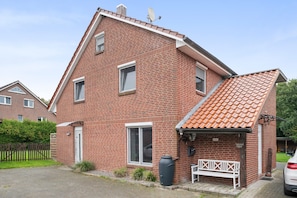 Een herenhuis met twee verdiepingen, een schuin dak, een voortuin met een bankje en een grote, zwarte waterreservoir.