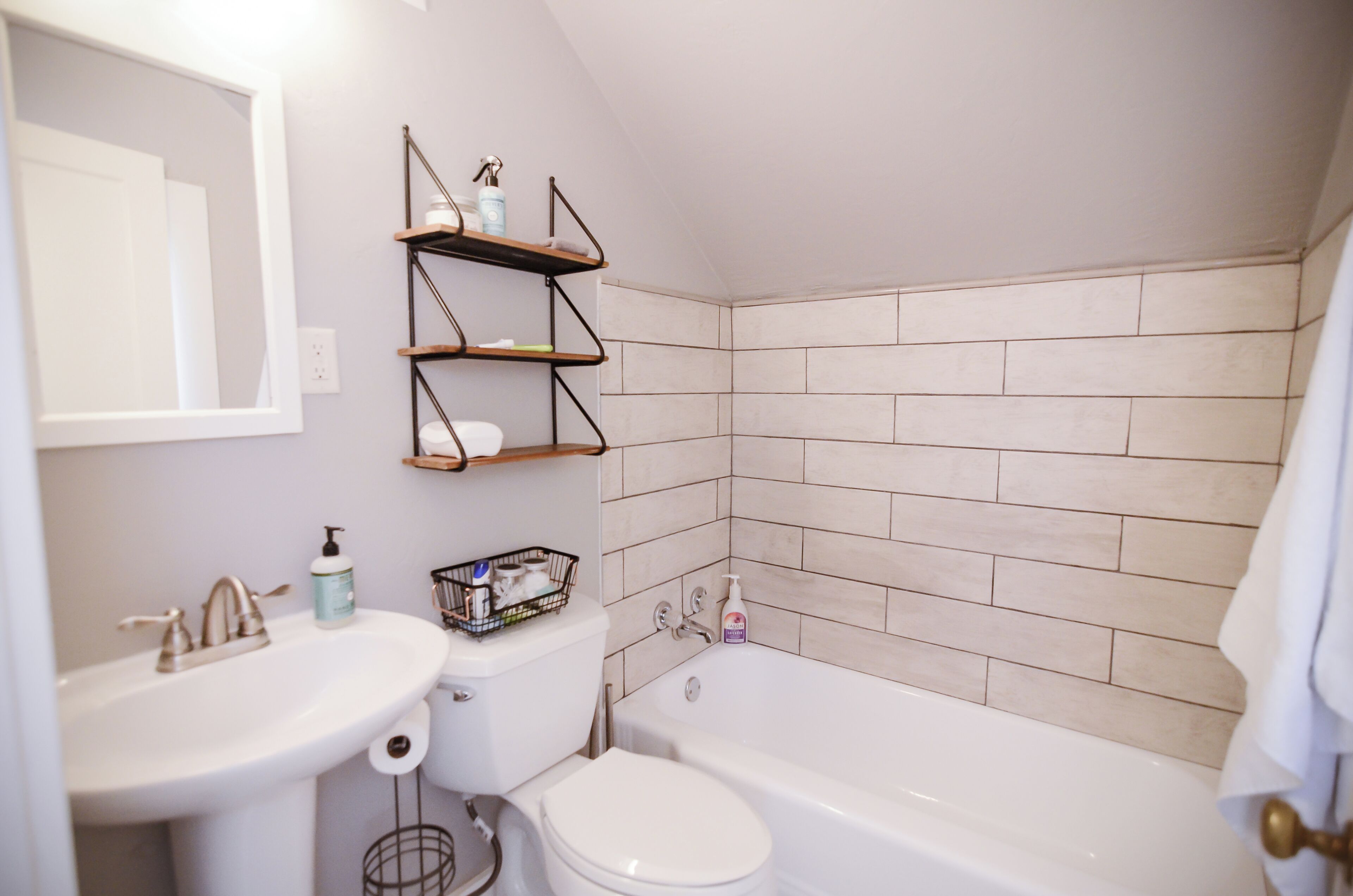 A bathroom with a sink, toilet, and bathtub, featuring tiled walls and shelves with toiletries.