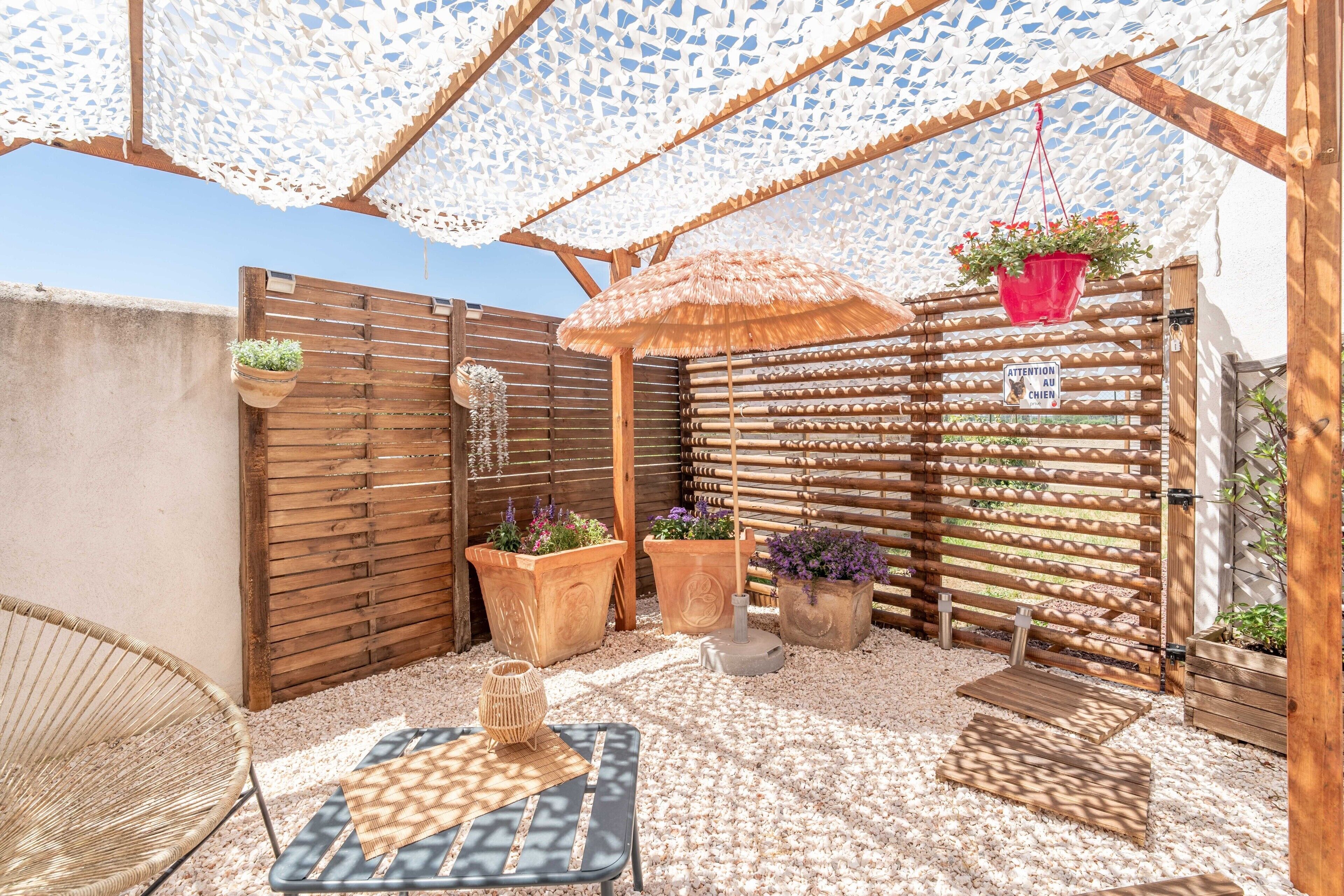 A patio with a woven chair, a table, potted plants, a wooden fence, and a thatched umbrella.