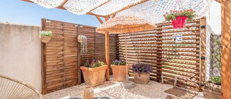 A patio with a woven chair, a table, potted plants, a wooden fence, and a thatched umbrella.