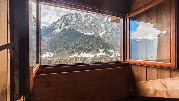 Una cabaña de madera con un ventanal grande que ofrece vistas a montañas nevadas y un pueblo en la falda.