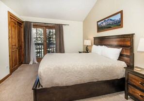 A bedroom with a large bed, wooden headboard, a nightstand with a lamp, a wooden wardrobe, and a window with curtains. - Modern Mountain East Vail Condo, Pool | TF1005 (Vail)