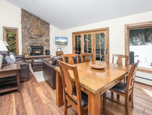 A cozy living room with a stone fireplace, a dining area with wooden furniture, and a view of a snowy landscape through large windows. - Modern Mountain East Vail Condo, Pool | TF1005 (Vail)