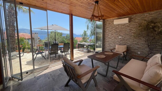 A modern outdoor patio with a view of the sea, a stone wall, and a wooden ceiling.