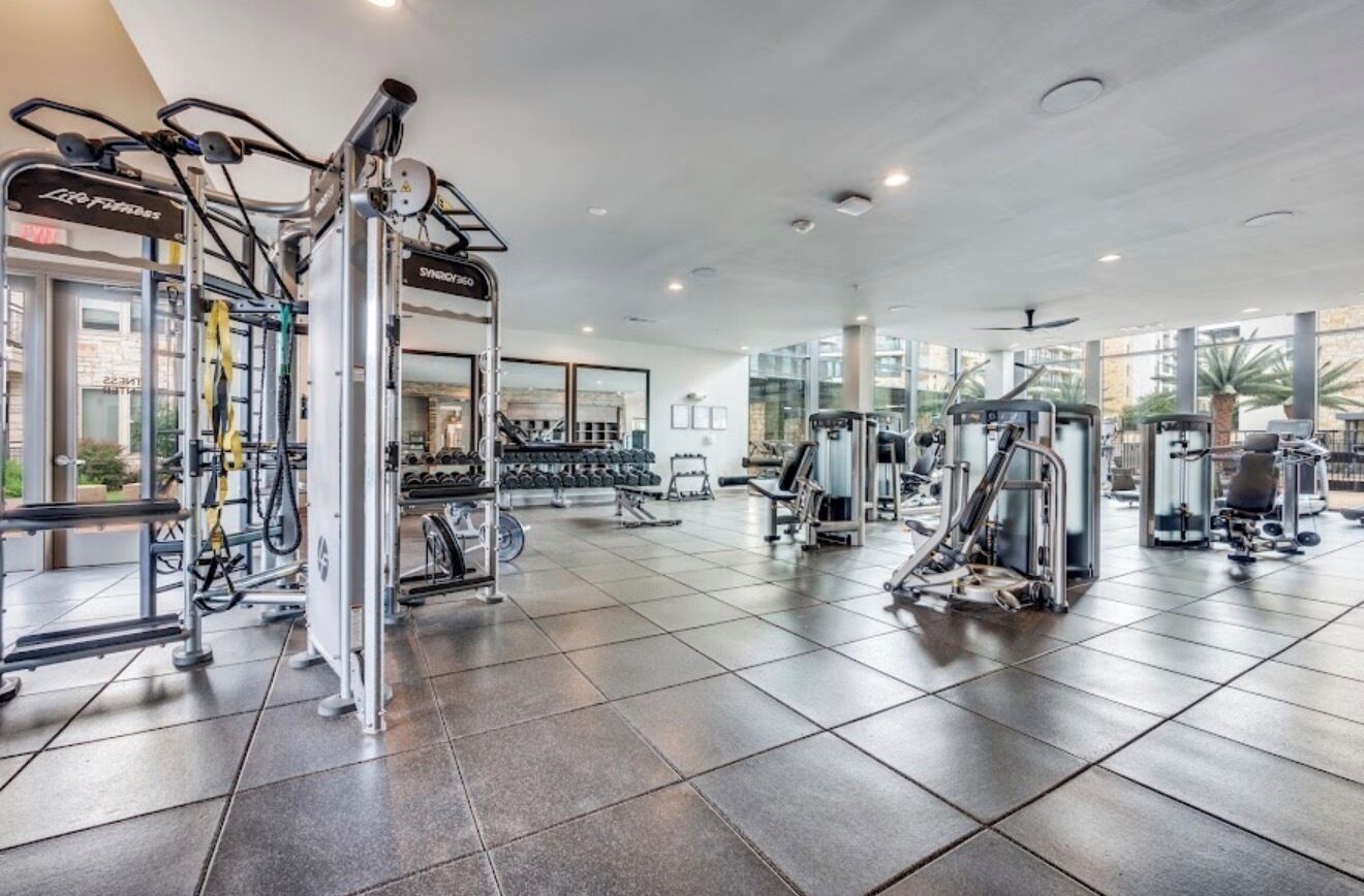 A gym with various exercise machines, a weight rack, and a large window providing natural light.