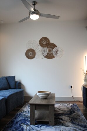 Een woonkamer met een plafondventilator, een blauwe bank, een bijzettafeltje met een schaal en wanddecoratie.