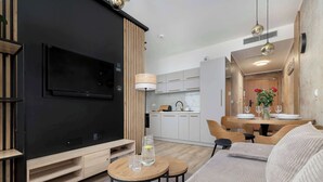 Ein modernes Wohnzimmer mit Flachbildfernseher, Holzelementen, einem Essbereich mit Tisch und Stühlen sowie einer Küche im Hintergrund.