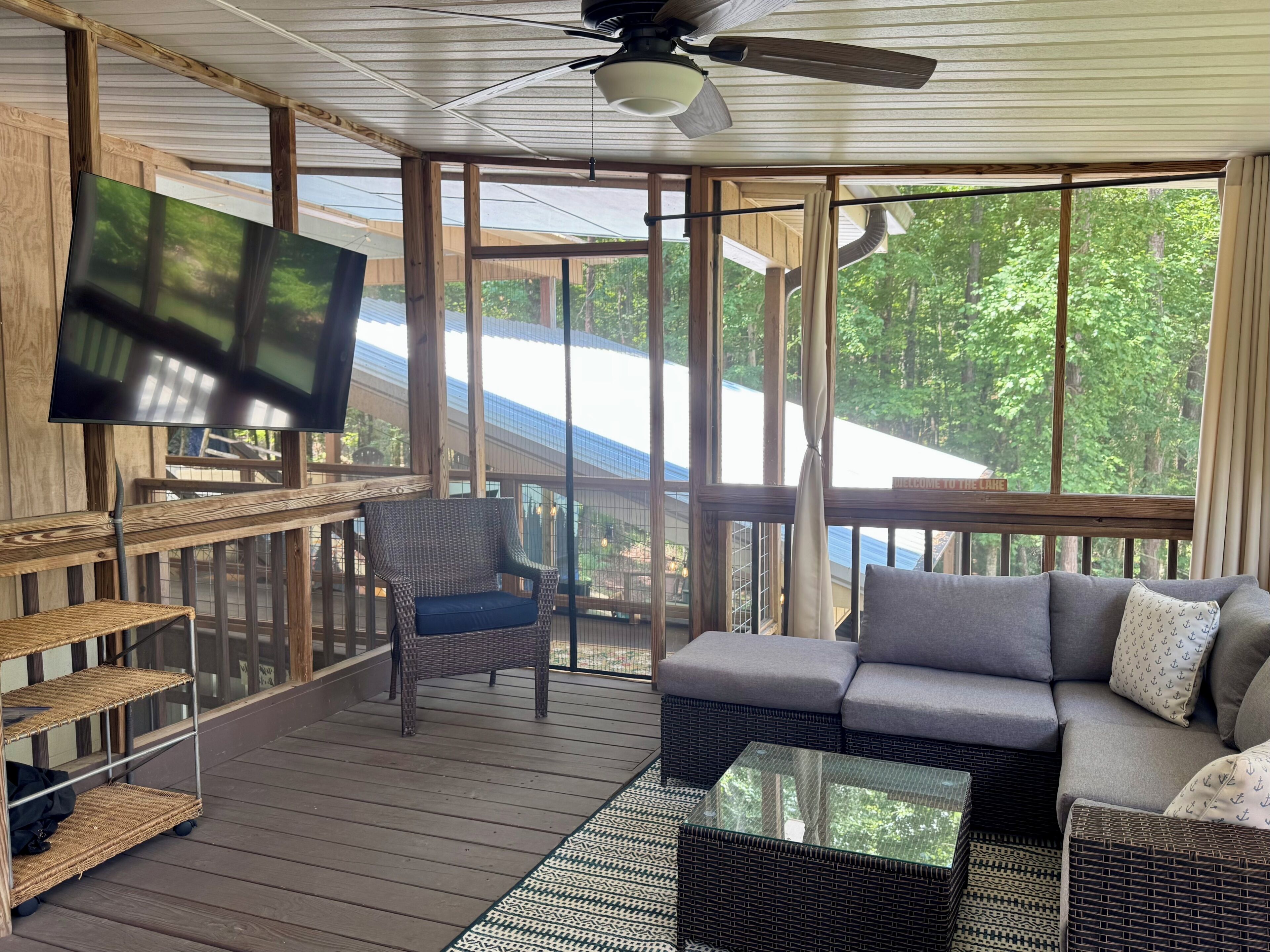 A covered outdoor area with a ceiling fan, a television, wicker furniture, and a view of trees through large windows.